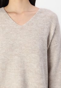 Beige gerippter Strickpullover mit V-Ausschnitt, weicher Textur und überschnittenen Schultern, der natürliche Variationen im Material zeigt.