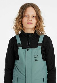 Salopette turquoise avec bretelles noires, dotée d'une poche avant et du logo "PRO TEST", portée avec une veste zippée noire en dessous.