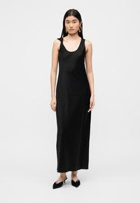 Robe maxi noire sans manches en tissu lisse ; présente un col dégagé et une silhouette ajustée. Le mannequin porte des escarpins noirs pointus.
