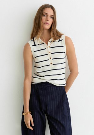 Donna con capelli lunghi che indossa una maglia bianca senza maniche con strisce orizzontali nere e pantaloni blu navy a righe, in piedi contro un muro bianco.