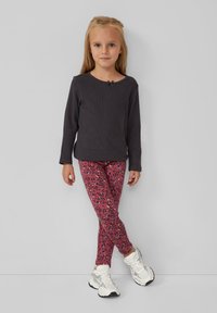 Młoda dziewczyna stoi przy ścianie, ubrana w ciemną bluzkę z długim rękawem, różowe legginsy w kwiatowy wzór i białe trampki.