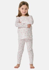 Ein junges Mädchen steht barfuß mit verschränkten Armen, trägt weiße Langarm-Pyjamas und eine Hose mit rosa Herzmustern.