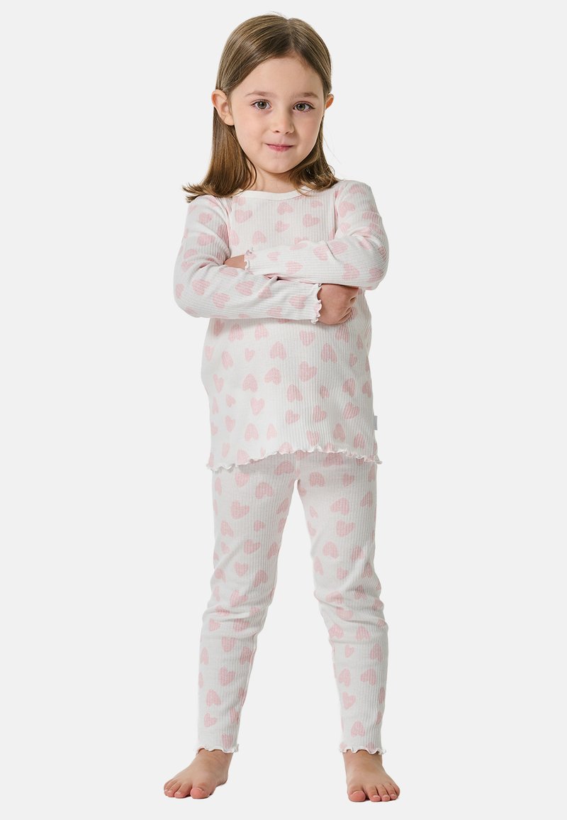 Ein junges Mädchen steht barfuß mit verschränkten Armen, trägt weiße Langarm-Pyjamas und eine Hose mit rosa Herzmustern.