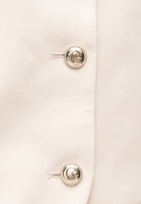 To sølvfarvede knapper med bogstavet "G" præget, fastgjort til lysebeige stof med synlige knaphuller.