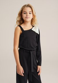 Djevojka s dugom plavom kosom, odjevena u crni kombinezon s jednom rukavom, s srebrnom svjetlucavom mašnom na ramenu, nasuprot bež pozadine.