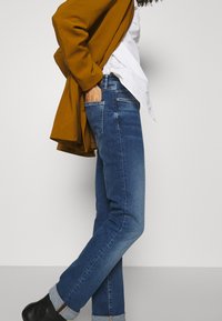 Džínové kalhoty v středně modré barvě se středním střihem. Mají ohrnuté manžety a jemný barevný přechod. Kombinují se s bílou košilí a hnědým svrchním oděvem.