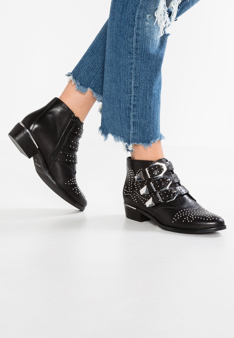 Botas de cuero negro hasta el tobillo con detalles de clavos plateados y tres correas ajustables con hebillas. Tacón bajo apilado, acabado texturizado y diseño de punta afilada.