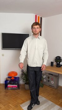 Camisa botão clara de cor cinza com um bolso no peito, jeans escuros e sapatilhas pretas. O fundo apresenta uma televisão e realces coloridos na parede.