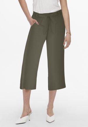 Mujer con pantalones anchos verde oliva recortados con cinturón delantero de lazo, top blanco sin mangas, mules blancos de tacón alto y pulsera dorada.