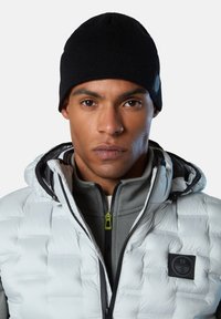 Cappello a maglia nero e giacca a piumino; la giacca è bianca con motivo in rilievo e dettagli neri; strato grigio con zip sotto, cursore della zip verde.