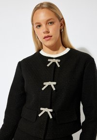 Svart texturerad jacka med tre dekorativa silverfärgad rosettknäppningar. Har en rund halsringning med kontrasterande vit kant och framfickor.