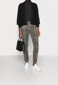 Černý pletený cardigan, černý rolák, vzorované béžovo-hnědé kalhoty s leopardím potiskem, bílé tenisky, držící malou černou kabelku.
