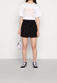 Weißes T-Shirt mit einem pinken grafischen Muster, kombiniert mit hochgeschnittenen schwarzen Shorts mit einer Frontbindeschleife. Schwarz-weiße Sneakers. Hellviolette Tasche.