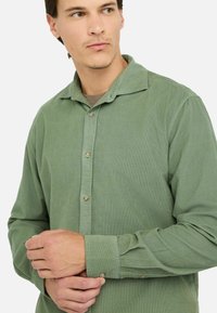 Camisa de pana verde claro con cierre de botones en la parte delantera, cuello extendido y mangas largas. Textura sutil con botones claros contrastantes.