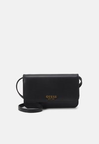Černá kožená crossbody taška s klopou, zlatým logem "GUESS EST 1981" a štíhlým popruhem. Hladká textura a minimalistický design.