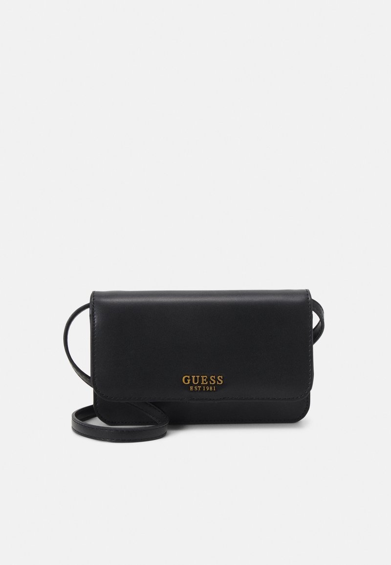 Černá kožená crossbody taška s klopou, zlatým logem "GUESS EST 1981" a štíhlým popruhem. Hladká textura a minimalistický design.