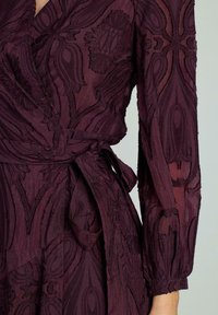 Rochie în nuanță de burgundy, din material transparent, cu broderie florală tonală intricate. Prezintă mâneci lungi, talie legată și accente texturate.