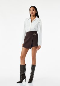 Witte langslavende blouse met kraag gecombineerd met een bruine wikkelrok met een gouden driehoekaccent. Model draagt zwarte kniehoge enkellaarsjes.