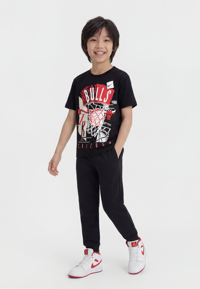 Ένα αγόρι που χαμογελά στέκεται, φορώντας μαύρο T-shirt με γραφικό των Chicago Bulls, μαύρα αθλητικά παντελόνια και λευκά με κόκκινα ψηλά αθλητικά παπούτσια Nike.