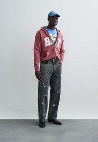 Röd zip-up hoodie med vitt tryck, blå grafisk t-shirt, grå jeans med broderade mönster, svarta skor och en blå keps.