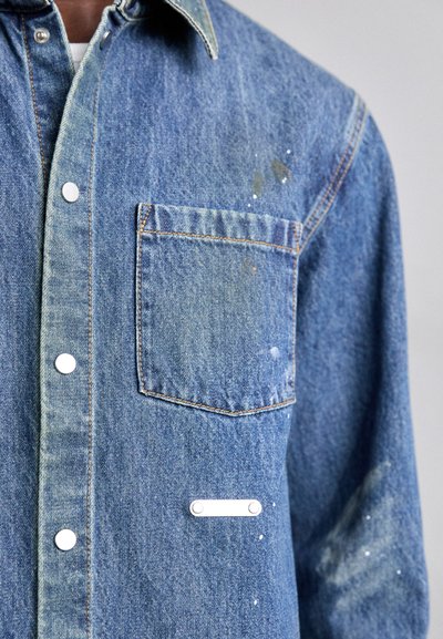 Camisa de denim en azul, con un bolsillo, botones plateados, una etiqueta de acento blanca y detalles de salpicaduras de pintura en la tela.