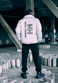 Witte pufferjack met zwart "Helly Hansen" logo en horizontale lijnen op de achterkant, gecombineerd met zwarte broek en schoenen. Schaars industrieel achtergrond.