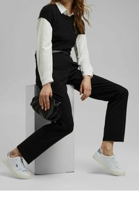 Femme portant un pantalon noir, une chemise blanche, un top sans manches noir, des baskets blanches, tenant une pochette noire tout en étant assise sur un bloc blanc.