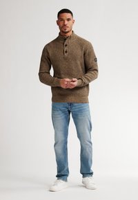 Man staan in een bruine gebreide trui met knopen, lichtblauwe jeans en witte sneakers tegen een effen achtergrond.