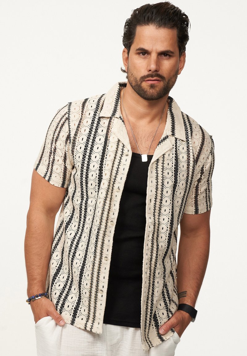 Kurzärmliges Hemd mit einem cremefarbenen Grund und schwarzen sowie beige geometrischen Muster, einem Kragen und Knopfverschluss, getragen über einem schwarzen Tanktop.