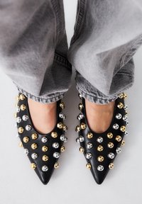 Mocasines negros de punta con adornos metálicos en dorado y plata, con un acabado de cuero suave y aberturas redondeadas.