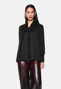 Schwarze Bluse mit langen Ärmeln, Kragen und Bindebanddetail, mit Falten und einer lässigen Silhouette, kombiniert mit burgunderfarbenen glänzenden Hosen.