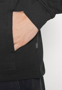 Camicia nera a maniche lunghe con tasca frontale, realizzata in tessuto liscio. Presenta una piccola etichetta con la scritta "FRIDAY" sulla cucitura laterale.