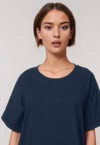 Una giovane donna con un taglio di capelli a bob corto indossa una t-shirt oversize di colore blu navy. Guarda direttamente verso la macchina fotografica su uno sfondo chiaro.