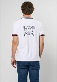 Hvid polo-shirt med et sort print featuring "135 År" og krydsede polo malletter; accentueret med marineblå og røde striber ved kraven og ærmerne.