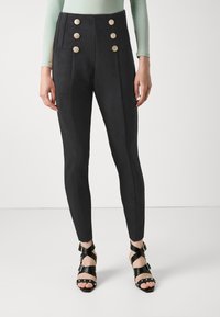 Pantaloni neri a vita alta con una texture liscia, caratterizzati da quattro bottoni decorativi dorati e tasche laterali, abbinati a tacchi neri con laccetti.
