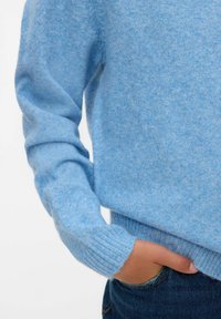 Ljusblå stickad tröja med ribbade ärmar och nederkant, som visar en mjuk textur och en avslappnad passform. Handen vilar försiktigt i jeans.