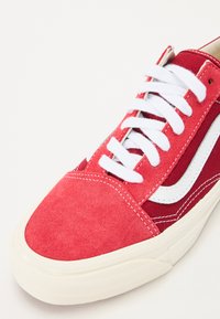 Red suede and canvas sneaker with white laces and stitching. Features a white side stripe and textured rubber sole. Close-up view.