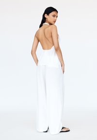 Top bianco senza schienale con design a collare halter, abbinato a pantaloni bianchi larghi. Il modello sta in posizione laterale, mettendo in mostra il tessuto e le linee semplici.