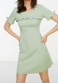 Kvinna i en ljusgrön kortärmad klänning med volangkant över bröstet och ärmar, stående med händerna på höfterna.