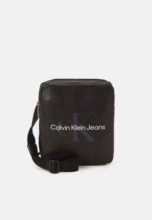 Bolso cruzado sintético negro con cierre de cremallera, que presenta un logo frontal que dice "Calvin Klein Jeans" en blanco, correa ajustable.