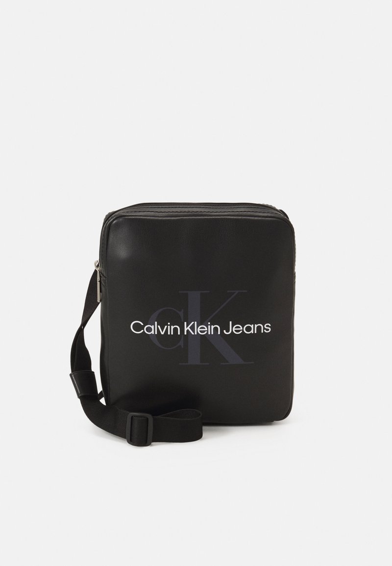 Borsa a tracolla sintetica nera con chiusura a zip, caratterizzata da un logo frontale che recita "Calvin Klein Jeans" in bianco, con tracolla regolabile.