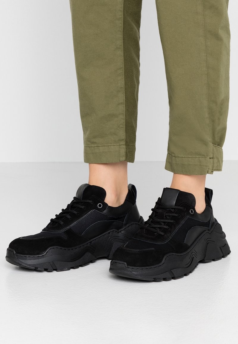 Baskets noires combinant des matériaux en suède et synthétiques, semelle épaisse, languette rembourrée et accents texturés, associées à un pantalon vert olive.