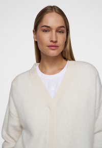 Crème-kleurige, zachte wollen cardigan met een V-hals ontwerp, verlaagde schouders en minimale details, gedragen over een wit shirt.