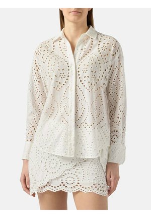 Donna che indossa una camicia bianca in pizzo sangallo a manica lunga con bottoni sul davanti e gonna corta abbinata con bordo smerlato.