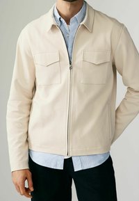 Veste beige zippée avec une finition texturée, deux poches poitrine et un col pointu, portée par-dessus une chemise rayée bleu clair et blanc.