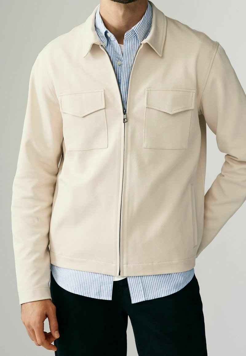 Veste beige zippée avec une finition texturée, deux poches poitrine et un col pointu, portée par-dessus une chemise rayée bleu clair et blanc.
