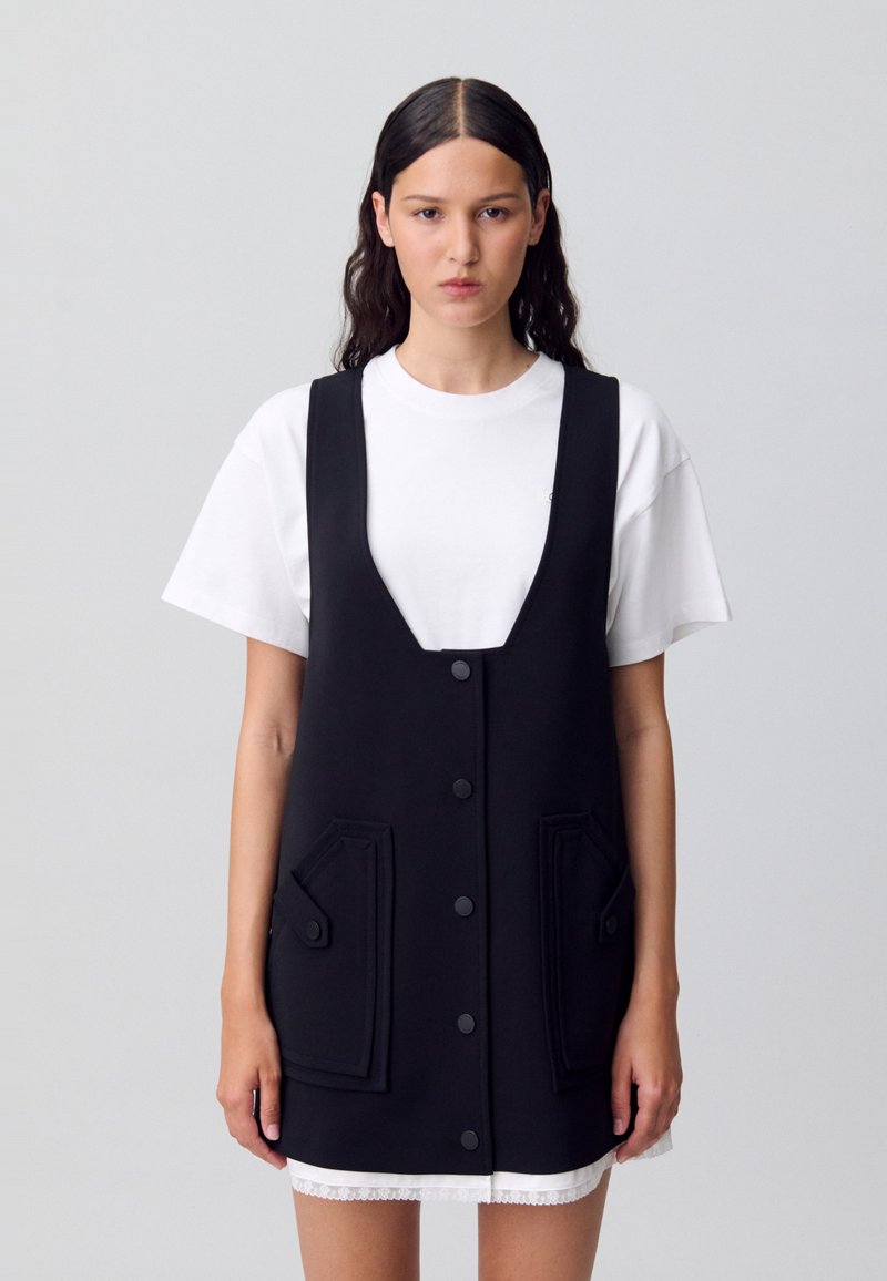 Abito nero senza maniche con scollatura a V, bottoni frontali, due tasche a bustina e una superficie liscia, indossato sopra una t-shirt bianca.