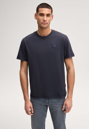 Mann mit kurzem Haar, der ein einfarbiges dunkelblaues T-Shirt und blaue Jeans trägt und vor einem hellgrauen Hintergrund steht.