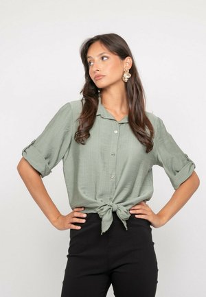 Femme aux longs cheveux portant une chemise verte à boutons nouée à la taille et un pantalon noir, les mains sur les hanches, regardant sur le côté devant un fond uni.