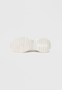 Talonul unui pantof sport alb, cu o suprafață din cauciuc texturat, având modele de talpă distincte pentru o aderență și stabilitate îmbunătățite.